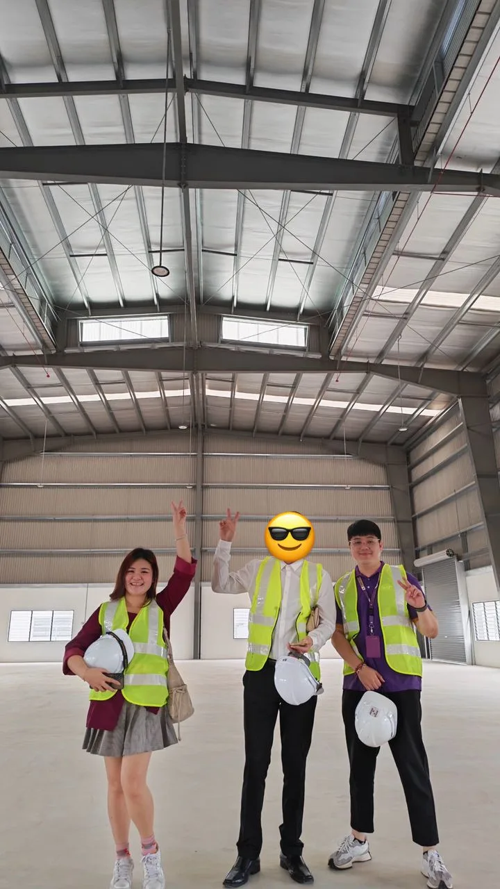 Three people wearing safety vests and holding hard hats inside a large industrial building.