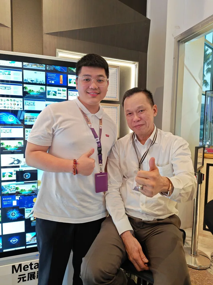 Two people giving thumbs up beside a wall of digital displays at an indoor event.