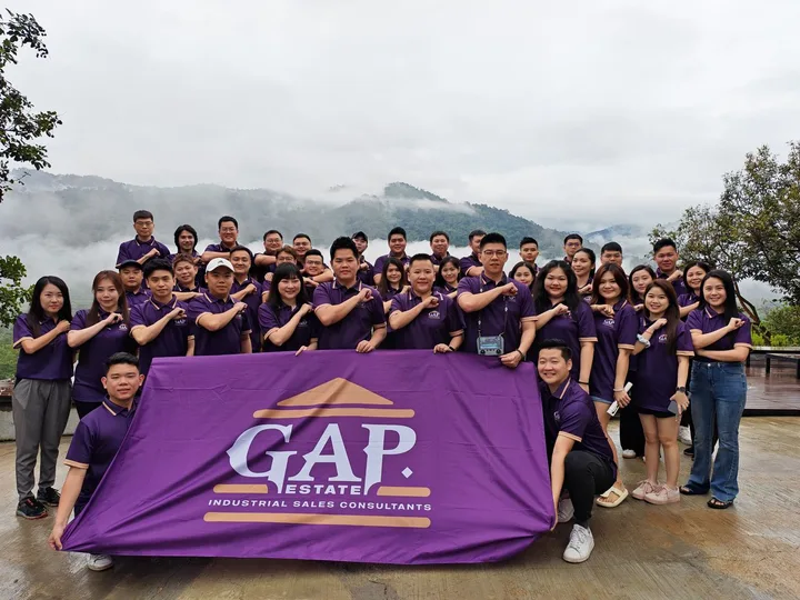 A large team in purple shirts posing outdoors with a banner and hills in the background.