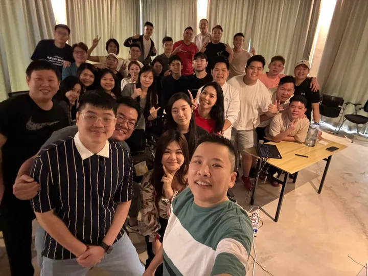 A large group gathering for a wide-angle selfie in a room with tables and chairs.